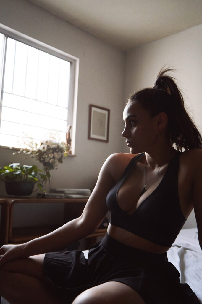 Valeria Cisneros a fitness enthusiast sits contemplatively in a sunlit room wearing a black sports bra and skirt with a necklace and earrings and a framed artwork on the wall behind her.